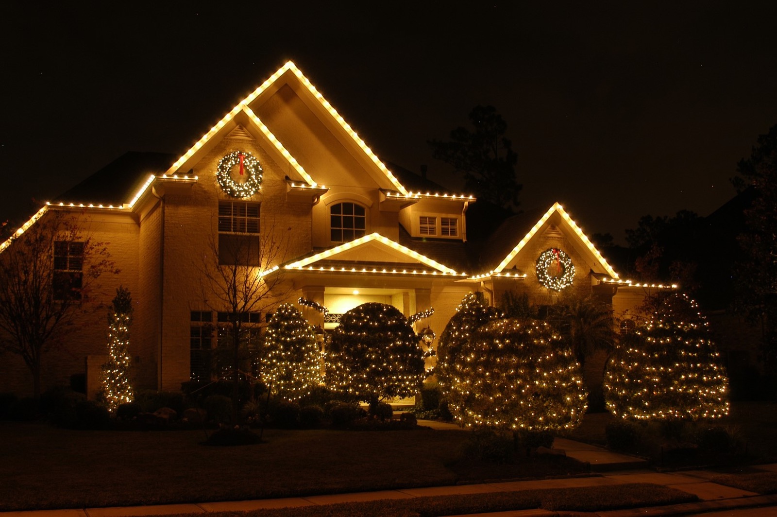 Warm white holiday lights outlining a home's roofline with wreaths and lit topiary shrubs